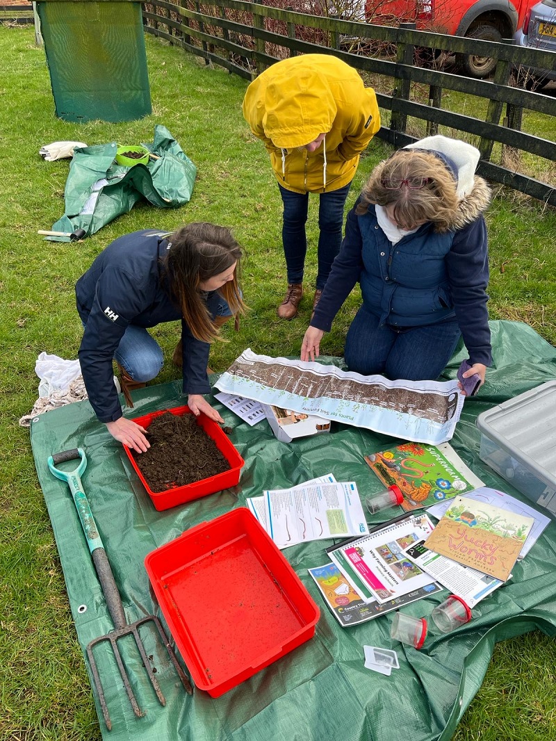 Three adults examining soil in red trays on a green tarpaulin 